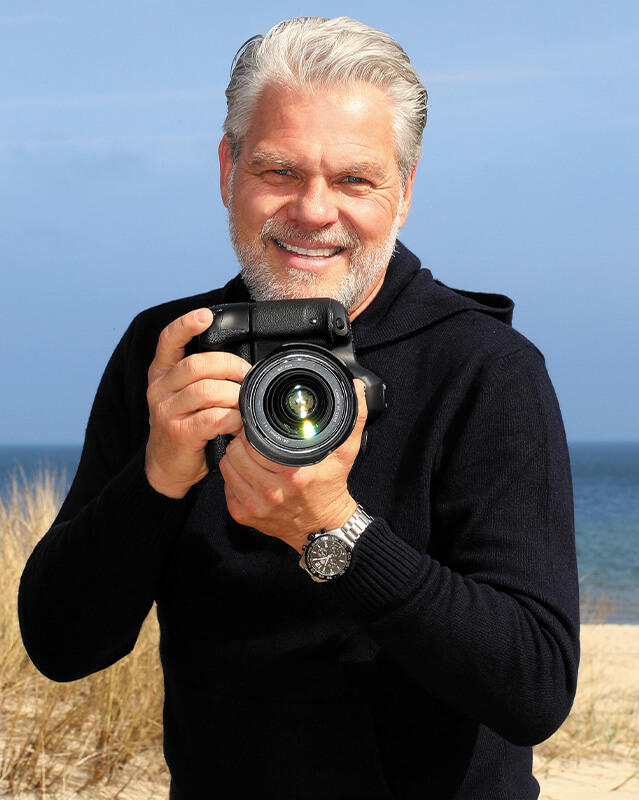 Fotograf mit Kamera in der Hand am Strand lächelnd
